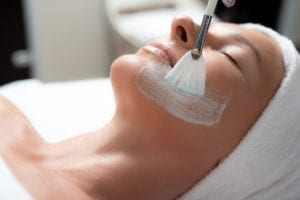 Close up portrait of beautiful middle aged woman with cosmetic clay for facial on her cheek and chin. She is lying on massage table with closed eyes and soft towel on head