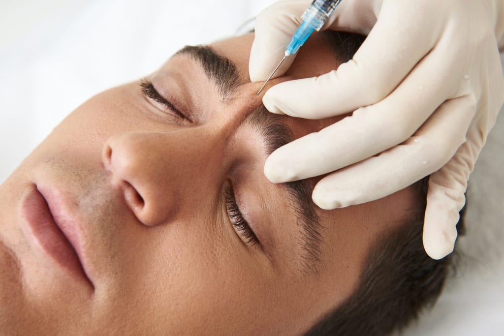 Close up top angle portrait of young male closing eyes while having in cosmetic center rejuvenating injections on forehead - Medical Spa Treatments